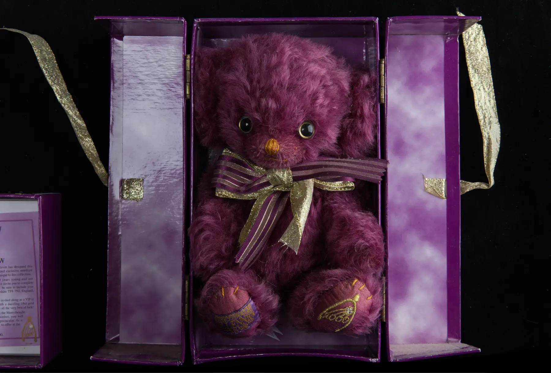 A deep magenta mohair teddy bear seated in an open purple presentation box with gold ribbons, a gold-tipped bow at its throat