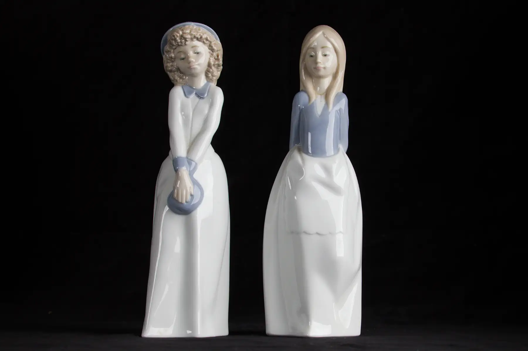 Two porcelain figurines of young women in long white gowns with pale blue bodices, one in a bonnet holding a hat, the other with long fair hair